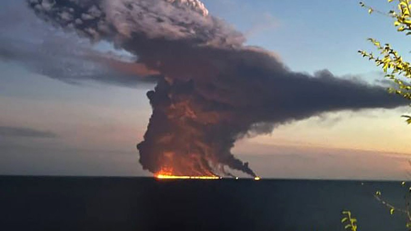 Fire line on the water, black smoke column, dusk sky, trees in the foreground.