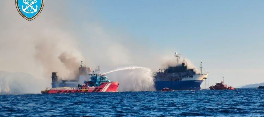 Landscape. Blue-hulled Ro-Ro, white-grey smoke over the after decks, a red-and-white tug spraying.