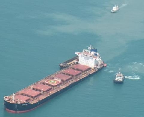 Black-hulled bulk carrier, KMAX EMPEROR on the port bow, tug at starboard aft, teal water.