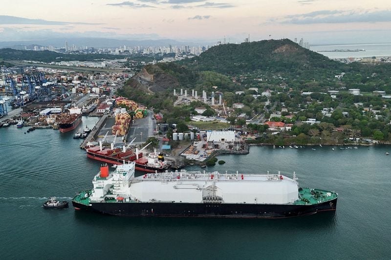 Aerial. Moss-type LNG carrier in the Panama Canal approaches, four spheres, tug at the stern, city haze beyond.