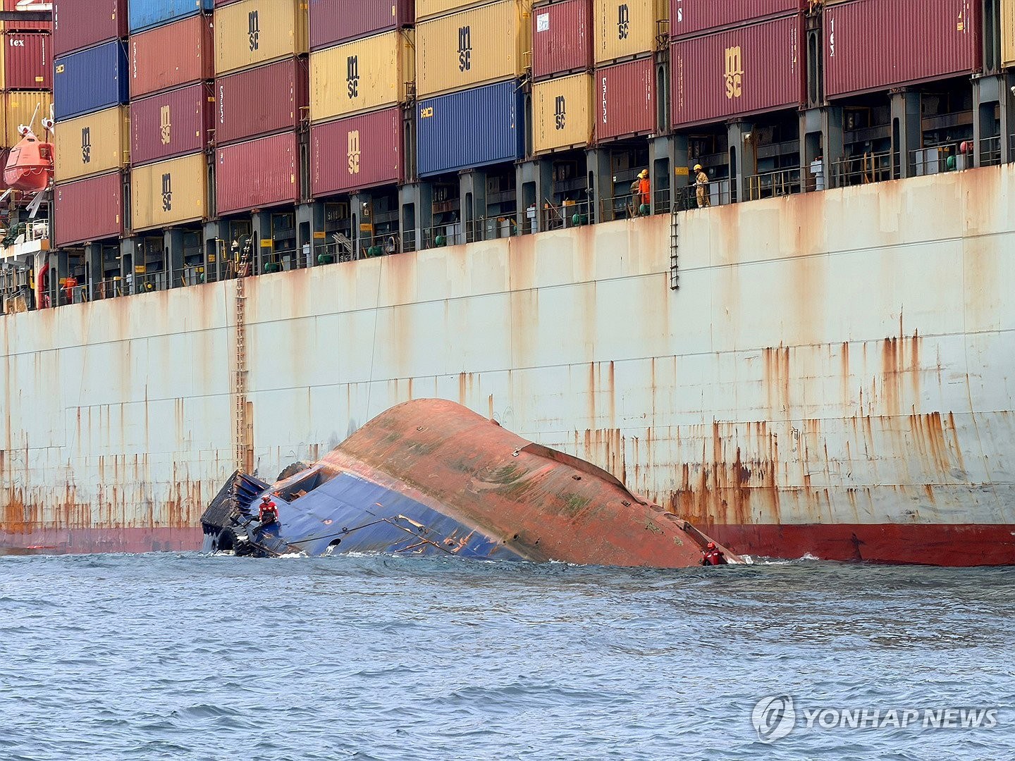 Capsized tug against a containership hull, figures on the upturned bottom.